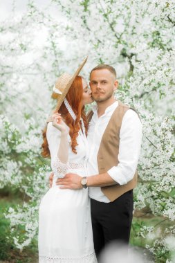 Beyaz çiçeklerin arka planında kucaklaşan sevgi dolu bir çift. Eski tarz, hasır şapkalı kız..