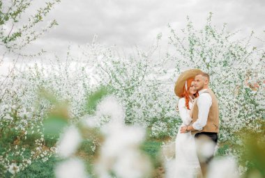 Beyaz çiçeklerin arka planında kucaklaşan sevgi dolu bir çift. Eski tarz, hasır şapkalı kız..