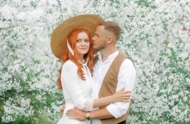 Beyaz çiçeklerin arka planında kucaklaşan sevgi dolu bir çift. Eski tarz, hasır şapkalı kız..