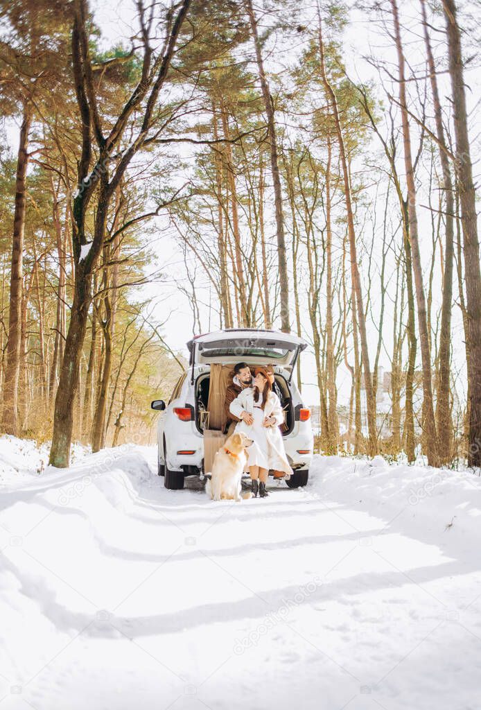 Smiling couple with beige dog sitting in open SUV car trunk in snowy forest. Enjoying each other in active winter holidays.