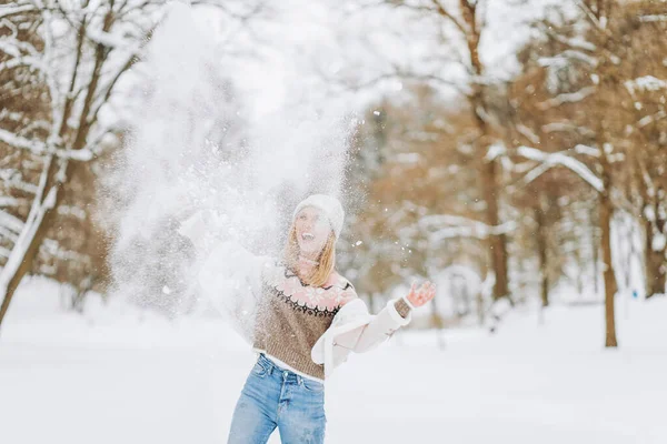 Mutlu komik sarışın kadın soğuk mevsimin keyfini çıkarmak için ellerini havaya kaldırarak kar fırlatıyor.