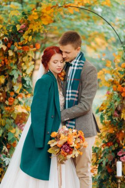 Mutlu yeni evliler sonbahar parkında düğün kemerinin yanında duruyor..