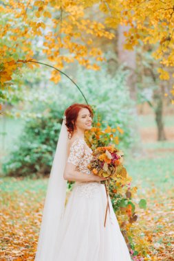 Gelinlik giymiş, elinde buketi olan ve sonbahar bahçesinin arka planında poz veren güzel kızıl saçlı kız..