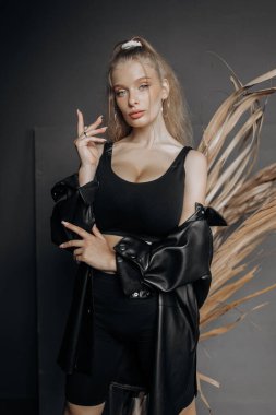 Şık bir model, sarı saçlı, siyah modaya uygun giyinmiş gri arka planda kuru otlarla süslenmiş poz veriyor..