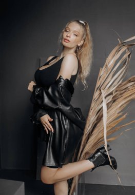 Şık bir model, sarı saçlı, siyah modaya uygun giyinmiş gri arka planda kuru otlarla süslenmiş poz veriyor..