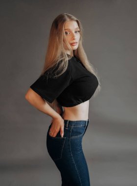 Kısa bluz ve kot pantolon giymiş, gri arka planda poz veren lüks bir sarışın.