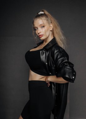 Sarı saçlı, şık giyinmiş, şort ve deri gömlekli, koyu bir arka planda poz veren güzel bir kız..