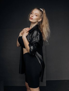 Sarı saçlı, şık giyinmiş, şort ve deri gömlekli, koyu bir arka planda poz veren kız..