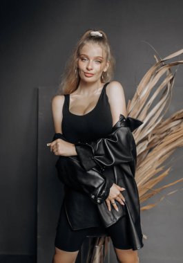 Şık bir model, sarı saçlı, siyah modaya uygun giyinmiş gri arka planda kuru otlarla süslenmiş poz veriyor..