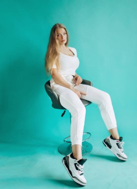 Stüdyoda sandalyede oturan beyaz takım elbiseli güzel sarışın. Turkuaz arkaplan.