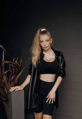 Güzel bir kız, sarı saçlı şık bir bluz, şort ve deri gömlekli koyu bir arka planda poz veriyor..