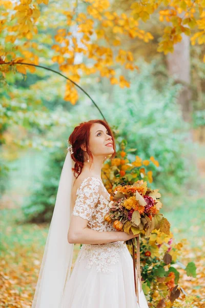 Gelinlik giymiş, elinde buketi olan ve sonbahar bahçesinin arka planında poz veren güzel kızıl saçlı kız..
