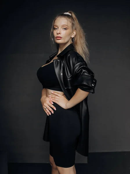 Güzel bir kız, sarı saçlı şık bir bluz, şort ve deri gömlekli koyu bir arka planda poz veriyor..