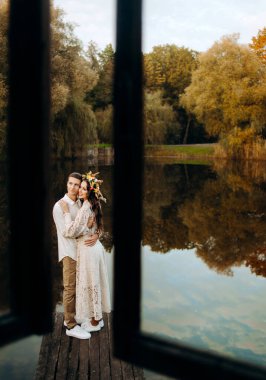Sevgi dolu bir çift, şenlikli giysiler içinde gölün arka planındaki iskelede duruyor, evin penceresinden bakıyor.