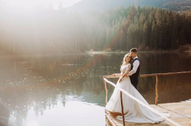 Genç çift birbirlerinin gözlerinin içine bakıyor, bir dağ gölünün yakınındaki iskelede el ele duruyorlar.