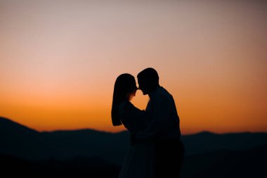 Damat ve gelin, gün batımının arka planında, dağda duruyorlar.