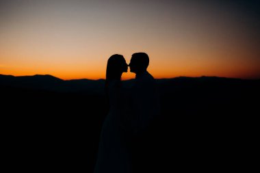 Damat ve gelin, gün batımının arka planında, dağda duruyorlar.