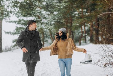 Genç bir çift donmuş bir gölün yakınındaki karlı bir parkta yürüyor. Kız elinde paten tutuyor..