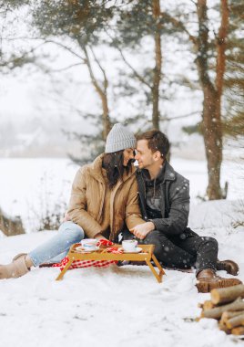 Karlı ormanda kış pikniği. Çam ağaçlarının altında oturmuş çay içen sevimli bir çift. Sevgililer Günü, romantizm.