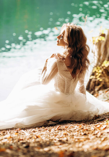 Luxury bride sitting near the lake in the sun, caressing her face.
