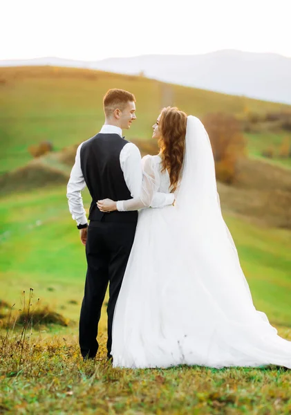 一对新婚夫妇穿着一件白色的婚纱和一套西服在山岗上走来走去 夕阳西下 — 图库照片