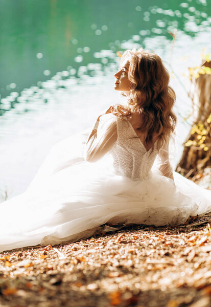 Amazing bride sitting near the lake in the sun, caressing her face.