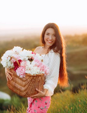 Güneş batarken poz veren, üzerinde beyaz ve pembe şakayık çiçekleri olan sepeti tutan mutlu kız. Yaşam tarzı, seyahat kavramı