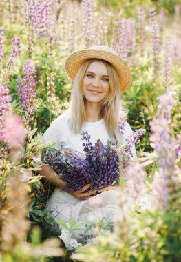 Hasır şapkalı çekici kız elinde bir buket lupinle bir tarlada oturuyor..