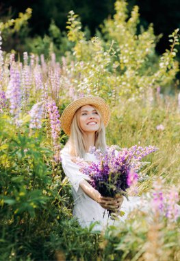 Hasır şapkalı çekici kız elinde bir buket lupinle bir tarlada oturuyor..