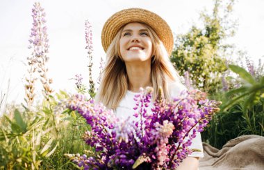 Hasır şapkalı çekici kız elinde bir buket lupinle bir tarlada oturuyor..