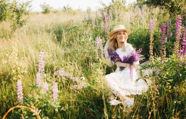 Hasır şapkalı çekici kız elinde bir buket lupinle bir tarlada oturuyor..
