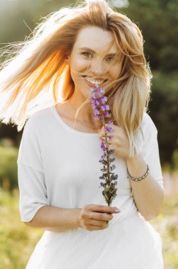 Rüzgâr genç bir sarışının saçlarını savuruyor elinde bir lup çiçeğiyle poz veriyor.