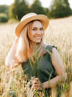 Hasır şapkalı, yeşil takım elbiseli, buğday tarlasında elinde çivilerle poz veren güzel bir sarışın.