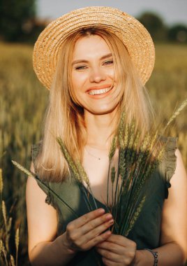 Hasır şapkalı, yeşil takım elbiseli, buğday tarlasında elinde çivilerle poz veren güzel bir sarışın.