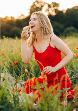 Kırmızı elbiseli güzel sarışın gün batımında çikolata parçacıklı kurabiye yiyor, gelincik tarlasında piknik yapıyor.