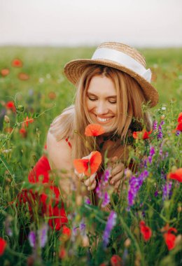 Hasır şapkalı ve kırmızı elbiseli mutlu sarışın tarlada haşhaş kokluyor..