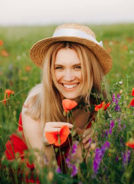 Hasır şapkalı ve kırmızı elbiseli mutlu sarışın tarlada haşhaş kokluyor..