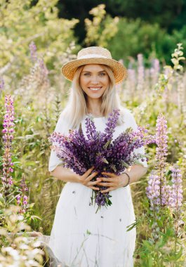 Hasır şapkalı çekici kız elinde bir buket lupinle bir tarlada oturuyor..