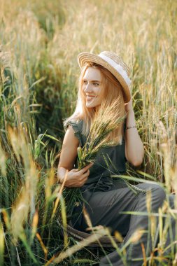 Hasır şapkalı güzel sarışın ve yeşil takım elbiseli buğday tarlasında oturuyor..