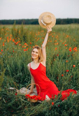 Çimlerin üzerinde oturan kırmızı elbiseli oyuncu sarışın. Gün batımında gelincik tarlasında piknik