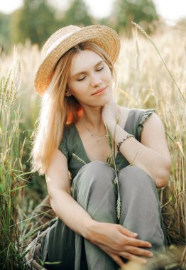 Hasır şapkalı güzel sarışın ve yeşil takım elbiseli buğday tarlasında oturuyor..