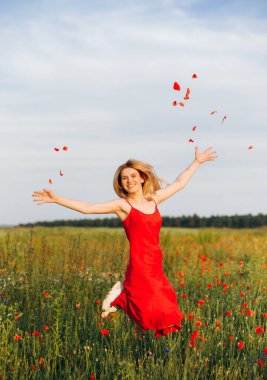 Kırmızı elbiseli zayıf sarışın gelincik tarlasına atlayıp gelincik yaprakları fırlatıyor.