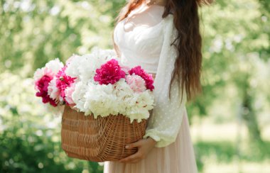 Saman sepetinde pembe şakayık tutan Boho kızı. Bohem çiçekli elbiseli şık hippi kadın bahçede şakayık çiçekleri topluyor..