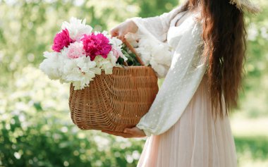 Saman sepetinde pembe şakayık tutan Boho kızı. Bohem çiçekli elbiseli şık hippi kadın bahçede şakayık çiçekleri topluyor..