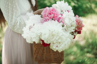 Saman sepetinde pembe şakayık tutan Boho kızı. Bohem çiçekli elbiseli şık hippi kadın bahçede şakayık çiçekleri topluyor..