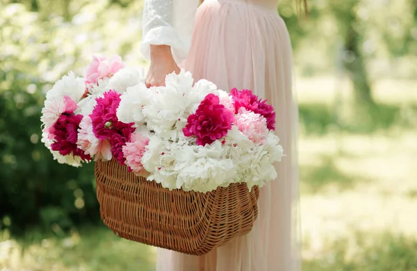 Boho女孩抱着粉红的牡丹在稻草篮 穿着波希米亚花裙子的时尚女人在花园里采摘牡丹花 — 图库照片