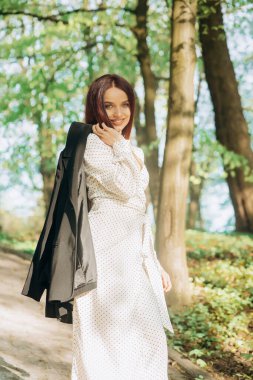 Beyaz elbiseli, siyah ceketli, güzel kızıl saçlı kız güneşli bir günde parkta yürüyor.