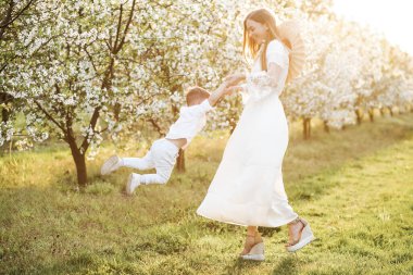 Anne oğlunun kollarını tutuyor ve etrafında dönüyor. Baharda çiçek açan bahçe.