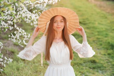Hasır şapkalı genç kadın baharda çiçek açan ağaçların bahçesinde poz veriyor..