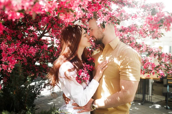 Genç mutlu, güler yüzlü bir çift Avrupa şehrinin caddesinde, pembe bahar çiçekleri yakınında poz veriyor..